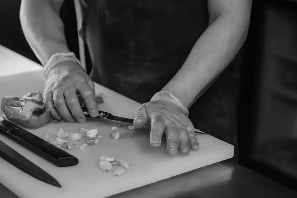A food preparer wearing disposable gloves chops garlic on a cutting board with a kitchen knife.