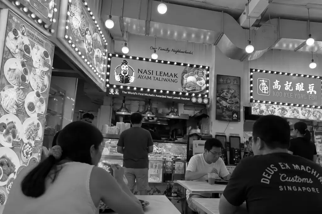 Diners having a meal at a Singapore food stall serving nasi lemak and other traditional dishes in a busy indoor food centre.