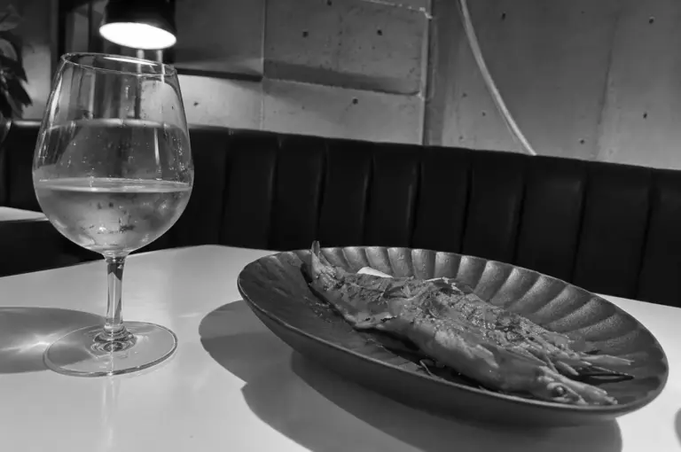 Side-angle close-up of a grilled prawn on a black plate beside a glass of white wine in a dimly lit restaurant.
