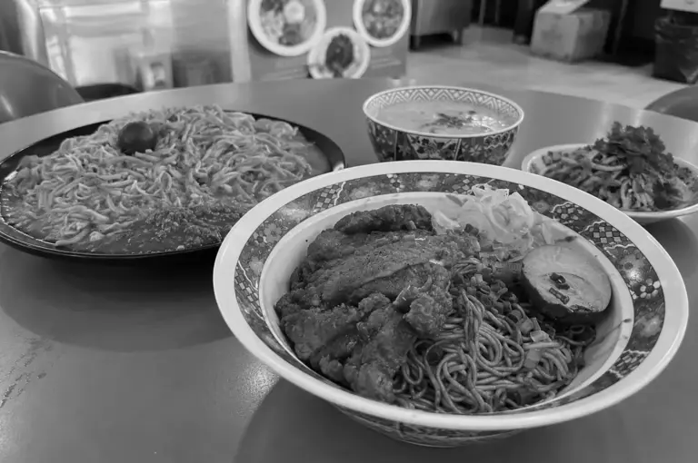 A bowl of Singapore-style noodles topped with sliced fish cake, braised meat, and leafy greens, served alongside other hawker noodle dishes on a shared table.