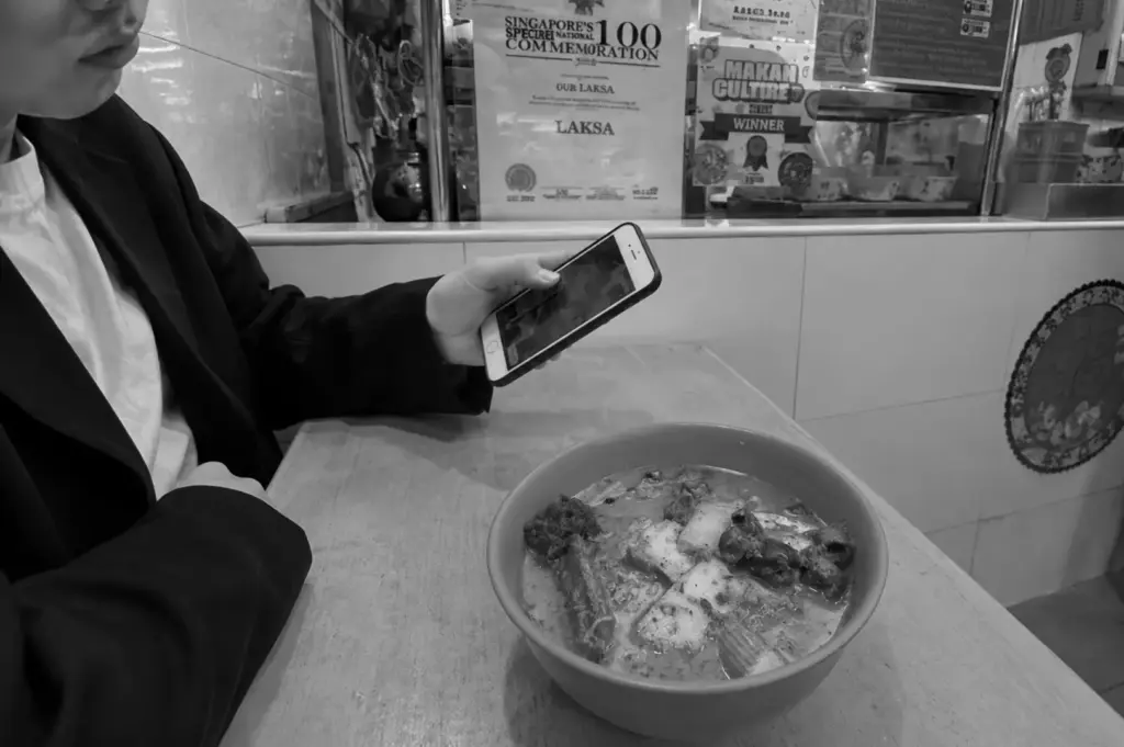 A customer photographing a bowl of traditional laksa with a smartphone inside a heritage food stall, showing how old-school food is being discovered through digital media.
