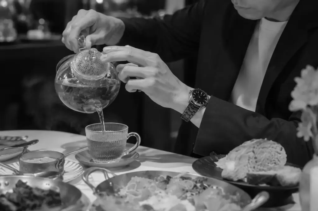 Close-up of a man pouring amber tea from a glass teapot into a textured glass cup at an elegant restaurant table.