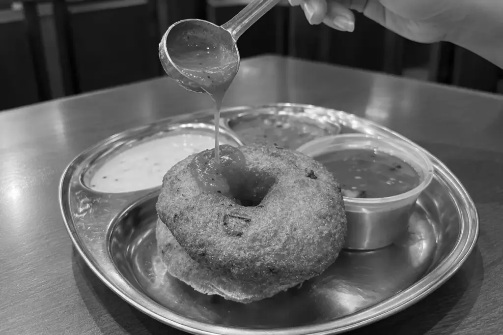 Medhu vadai served with chutney and sambar, with sauce being poured over the crispy savoury doughnut.