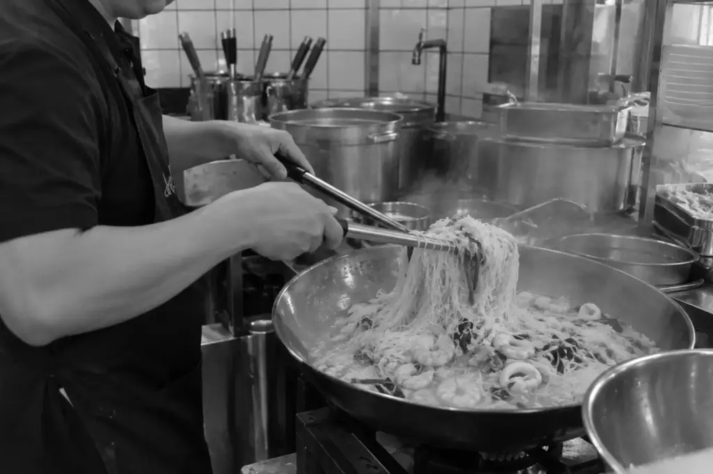 A chef tossing noodles with seafood in a large pan inside a working restaurant kitchen.